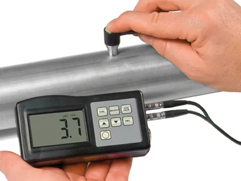 A handheld ultrasonic thickness meter reading a steel pipe, its probe held against the pipe wall